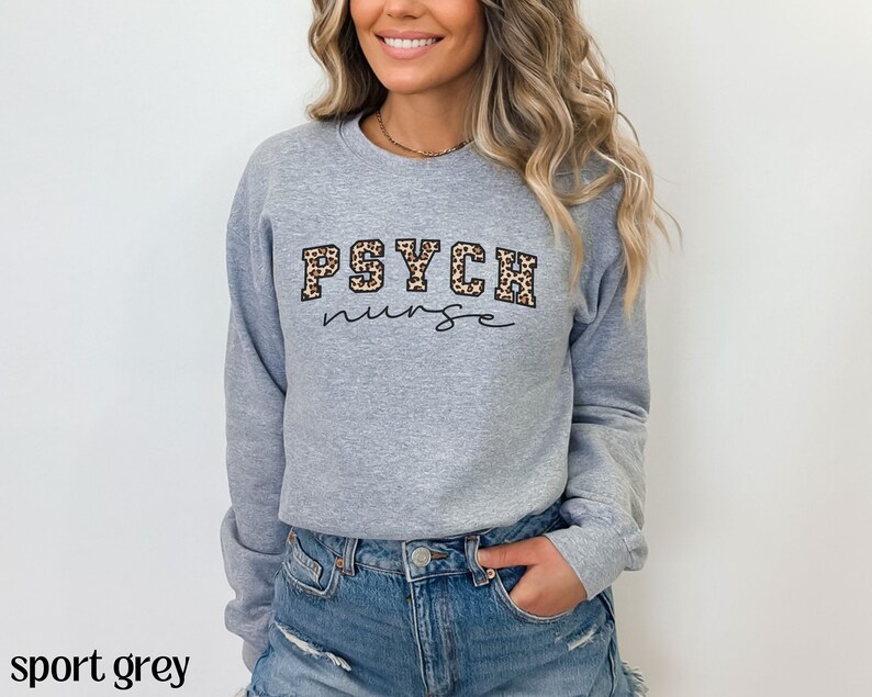 May include: A light grey sweatshirt with the text "PSYCH nurse" in a combination of leopard print and script font. The sweatshirt is a crew neck style and has long sleeves. The words "sport grey" are at the bottom of the image.