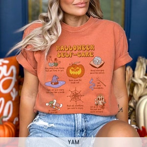 May include: Orange t-shirt with a Halloween self-care graphic. The graphic features a pumpkin, a witch's hat, a spiderweb, a potion bottle, a crystal ball, a tombstone, and candy. The text on the shirt reads "Halloween Self-Care".