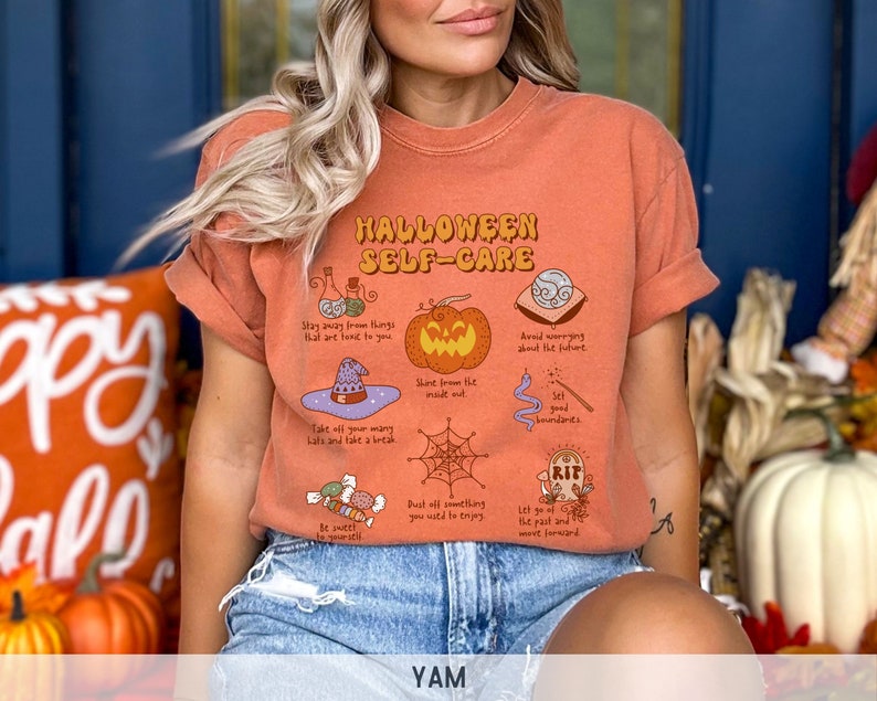 May include: Orange t-shirt with a Halloween self-care graphic. The graphic features a pumpkin, a witch's hat, a spiderweb, a potion bottle, a crystal ball, a tombstone, and candy. The text on the shirt reads "Halloween Self-Care".