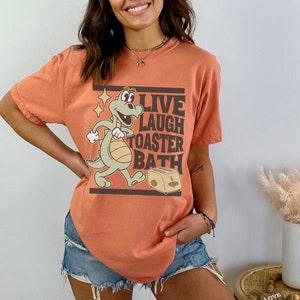 May include: Orange t-shirt with a cartoon dinosaur graphic and the text "LIVE LAUGH TOASTER BATH". The dinosaur is light green and tan, with a smiling expression. The toaster is tan. The shirt is a relaxed fit.