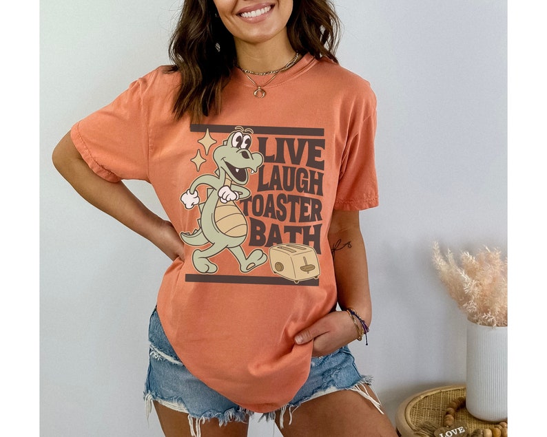 May include: Orange t-shirt with a cartoon dinosaur graphic and the text "LIVE LAUGH TOASTER BATH". The dinosaur is light green and tan, with a smiling expression. The toaster is tan. The shirt is a relaxed fit.