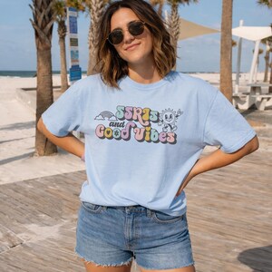 May include: Light blue t-shirt with the text "SSR's and Good Vibes" in a retro font. The graphic includes a rainbow, sun, and other elements. The shirt is paired with denim shorts.