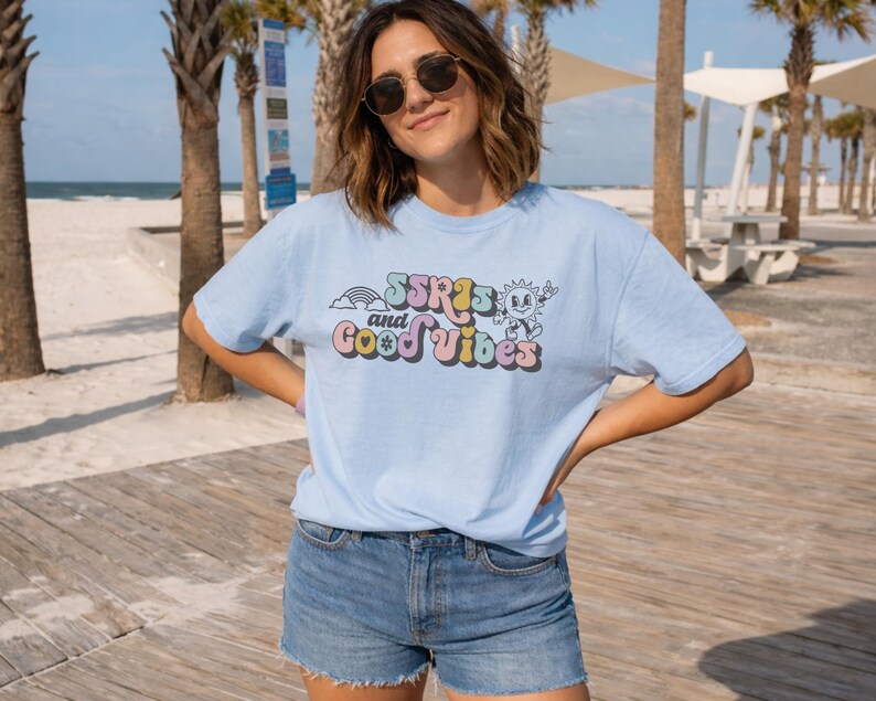 May include: Light blue t-shirt with the text "SSR's and Good Vibes" in a retro font. The graphic includes a rainbow, sun, and other elements. The shirt is paired with denim shorts.