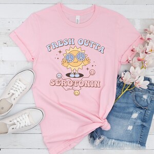 Op de afbeelding: Roze t-shirt met de tekst "FRESH OUTTA SEROTONIN" en een cartoon zon met een peace-teken zonnebril. Het shirt is gestyled met een spijkerbroek en witte sneakers op een wit houten oppervlak.