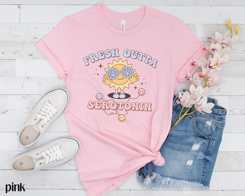 Op de afbeelding: Roze t-shirt met de tekst "FRESH OUTTA SEROTONIN" en een cartoon zon met een peace-teken zonnebril. Het shirt is gestyled met een spijkerbroek en witte sneakers op een wit houten oppervlak.