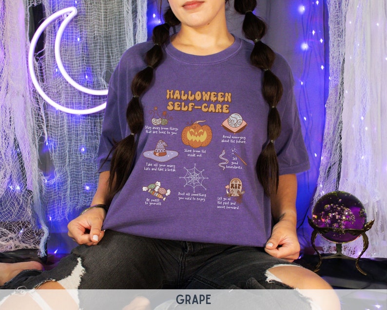May include: A purple t-shirt with a graphic print of a list of Halloween self-care tips. The text on the shirt reads "Halloween Self-Care".