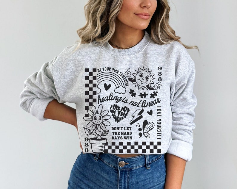 May include: Gray sweatshirt with a black and white graphic print featuring a flower, a sun, a rainbow, a heart, a lightning bolt, a butterfly, and the text "BE YOUR OWN SUNSHINE", "healing is not linear", "FIND JOY IN THE JOURNEY", "DON'T LET THE HARD DAYS WIN", "LOVE YOURSELF", and "988".