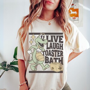 May include: Cream-colored t-shirt featuring a cartoon alligator with the text "LIVE LAUGH TOASTER BATH" in a black rectangular frame. The alligator is green and yellow, and the toaster is beige.
