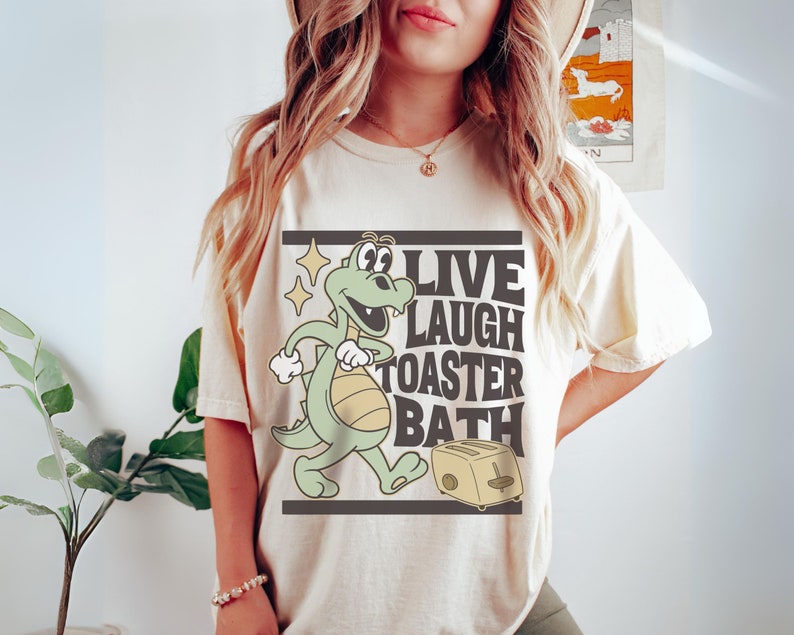 May include: Cream-colored t-shirt featuring a cartoon alligator with the text "LIVE LAUGH TOASTER BATH" in a black rectangular frame. The alligator is green and yellow, and the toaster is beige.