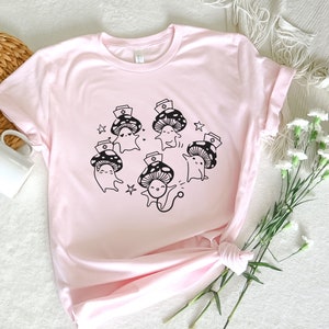 Op de afbeelding: Roze T-shirt met een afbeelding van vijf cartoon-paddenstoelen die verpleegstersmutsen dragen. De paddenstoelen glimlachen en hebben zwarte contouren. Het shirt heeft een ronde hals en korte mouwen.