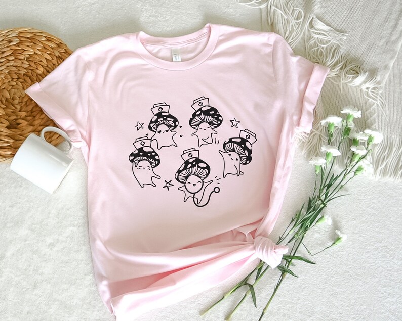 Op de afbeelding: Roze T-shirt met een afbeelding van vijf cartoon-paddenstoelen die verpleegstersmutsen dragen. De paddenstoelen glimlachen en hebben zwarte contouren. Het shirt heeft een ronde hals en korte mouwen.
