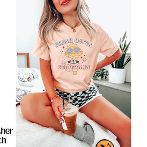 Op de afbeelding: Perzikkleurig T-shirt met de tekst "FRESH OUTTA SEROTONIN" en een cartoonzon met een vredestekenbril. De zon lacht en heeft een vredesteken. Het shirt heeft korte mouwen.