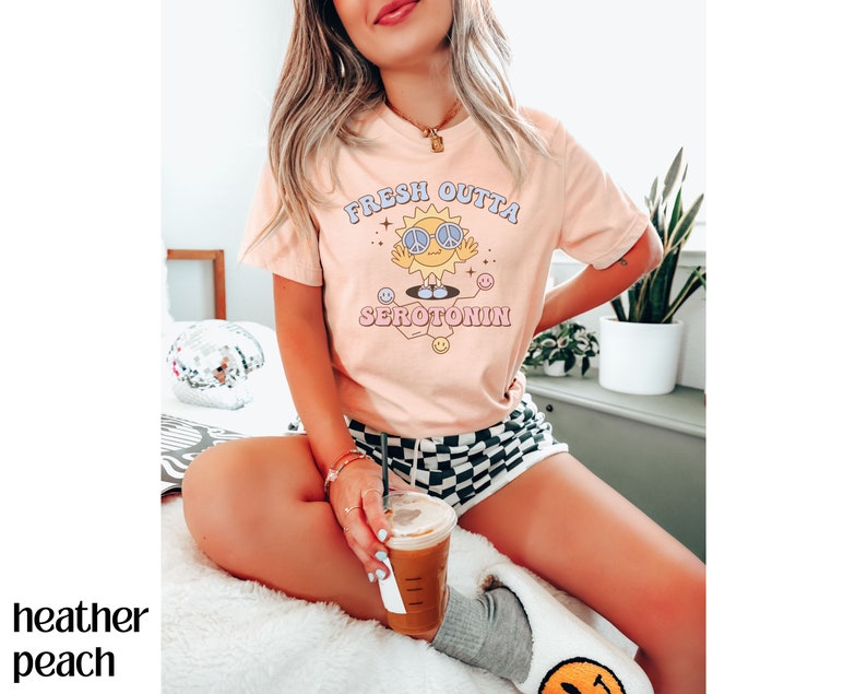 Op de afbeelding: Perzikkleurig T-shirt met de tekst "FRESH OUTTA SEROTONIN" en een cartoonzon met een vredestekenbril. De zon lacht en heeft een vredesteken. Het shirt heeft korte mouwen.