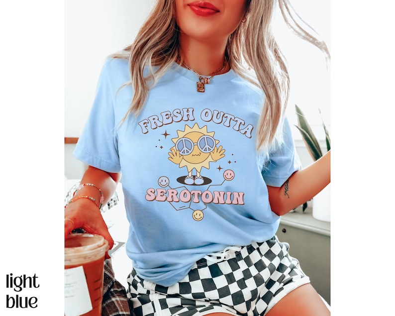 Op de afbeelding: Lichtblauw T-shirt met de tekst "FRESH OUTTA SEROTONIN" en een cartoon zon met een peace-teken bril. De zon heeft een lachend gezicht en is omringd door sterren en smiley gezichten. Het shirt heeft een lichtblauwe kleur.