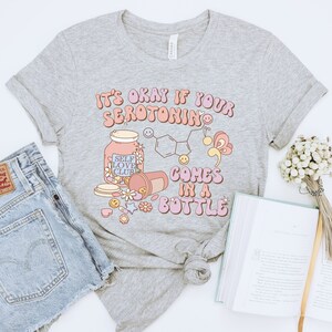 May include: Gray t-shirt with the text "It's okay if your serotonin comes in a bottle" in pink and orange letters. The design includes a pill bottle, a jar labeled "Self Love Club", and a chemical structure.