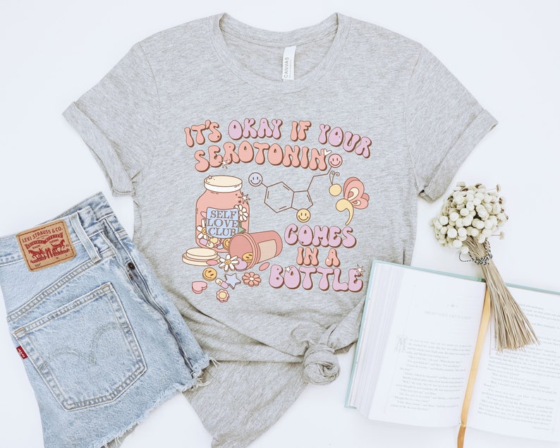May include: Gray t-shirt with the text "It's okay if your serotonin comes in a bottle" in pink and orange letters. The design includes a pill bottle, a jar labeled "Self Love Club", and a chemical structure.