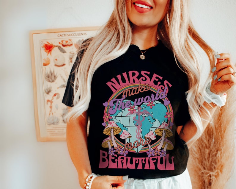 Puede incluir: Camiseta negra con un gr&aacute;fico de arco&iacute;ris y globo terr&aacute;queo. El texto "Nurses make the world more beautiful" est&aacute; impreso en la camiseta.