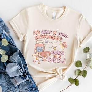 May include: A cream colored t-shirt with the text "It's okay if your serotonin comes in a bottle" in pink, purple, and blue lettering. The shirt features a cartoon illustration of a pill bottle with the text "Self Love Club" and a chemical structure of serotonin.