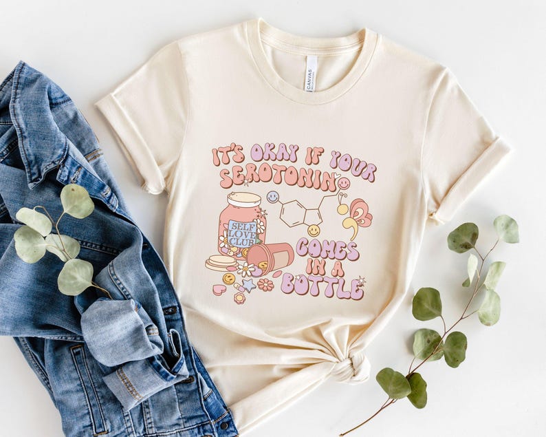 May include: A cream colored t-shirt with the text "It's okay if your serotonin comes in a bottle" in pink, purple, and blue lettering. The shirt features a cartoon illustration of a pill bottle with the text "Self Love Club" and a chemical structure of serotonin.