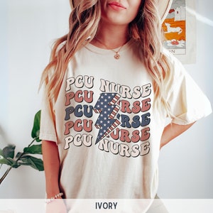 K&ouml;nnte beinhalten: Ein wei&szlig;es T-Shirt mit einem Grafikdruck eines Blitzes mit einem amerikanischen Flaggen-Design. Der Text "PCU NURSE" wird in einer Retro-Schriftart um den Blitz herum wiederholt. Das Shirt ist mit "IVORY" gekennzeichnet.