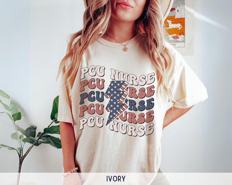 K&ouml;nnte beinhalten: Ein wei&szlig;es T-Shirt mit einem Grafikdruck eines Blitzes mit einem amerikanischen Flaggen-Design. Der Text "PCU NURSE" wird in einer Retro-Schriftart um den Blitz herum wiederholt. Das Shirt ist mit "IVORY" gekennzeichnet.