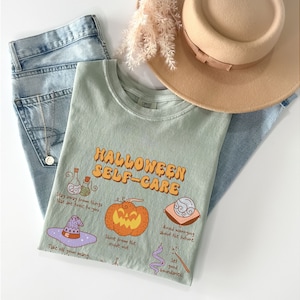 May include: A light green t-shirt with the text "HALLOWEEN SELF-CARE" in orange and yellow letters. The shirt features illustrations of a potion bottle, a pumpkin, a witch's hat, a crescent moon, and a snake with a wand. The text on the shirt reads: "Stay away from things that are toxic to you", "Shine from the inside out", "Avoid worrying about the future", and "Set good boundaries".