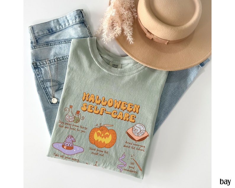 May include: A light green t-shirt with the text "HALLOWEEN SELF-CARE" in orange and yellow letters. The shirt features illustrations of a potion bottle, a pumpkin, a witch's hat, a crescent moon, and a snake with a wand. The text on the shirt reads: "Stay away from things that are toxic to you", "Shine from the inside out", "Avoid worrying about the future", and "Set good boundaries".