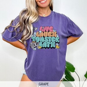 Pu&ograve; includere: Una maglietta viola con un design grafico che dice "Live Much Toaster Bath" in lettere a bolle colorate. La grafica include anche un tostapane dei cartoni animati e paperelle di gomma.