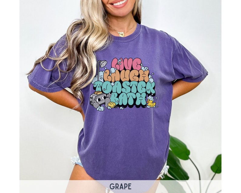 Pu&ograve; includere: Una maglietta viola con un design grafico che dice "Live Much Toaster Bath" in lettere a bolle colorate. La grafica include anche un tostapane dei cartoni animati e paperelle di gomma.