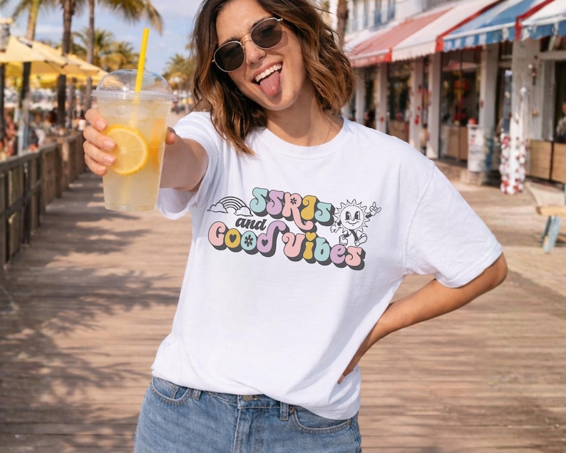 May include: White t-shirt with the text "SSRIs and Good Vibes" in a retro font, featuring a rainbow and a smiling sun graphic. The person is holding a clear plastic cup with a yellow straw and a lemon slice.