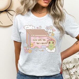 May include: A gray t-shirt with a graphic of a frog wearing a mushroom hat and a cloud with the text "Close some tabs take a breath". The graphic is surrounded by flowers and stars.
