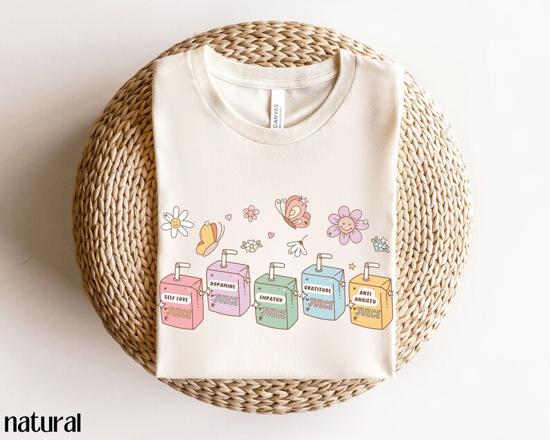 May include: A beige t-shirt with a graphic of colorful juice boxes with positive affirmations like "Self Love Juice", "Dopamine Juice", "Empathy Juice", "Gratitude Juice", and "Anti Anxiety Juice". The graphic also includes butterflies and flowers.