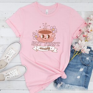 May include: Pink t-shirt with a cartoon illustration of a coffee cup with eyes and the text "But first, anxiety".