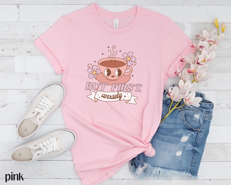 May include: Pink t-shirt with a cartoon illustration of a coffee cup with eyes and the text "But first, anxiety".