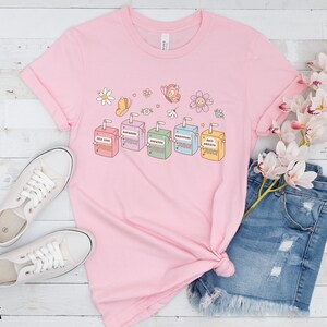 May include: Pink t-shirt with a graphic of colorful juice boxes with text that reads "Self Love Punch", "Empathy Punch", "Gratitude Punch", "Optimism Punch", and "Anti-Anxiety Punch".