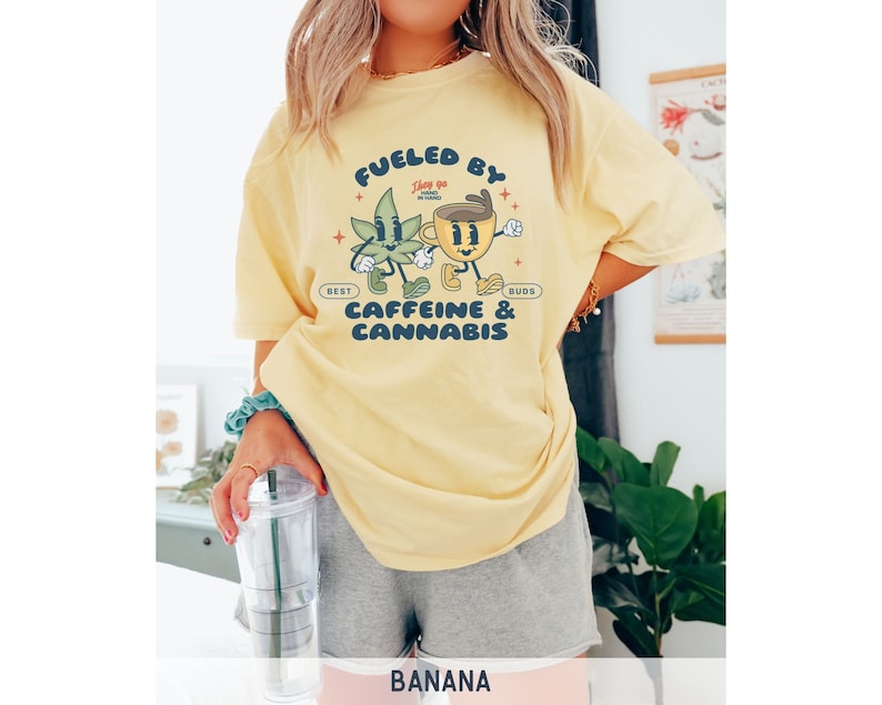 Puede incluir: Camiseta amarilla con un gr&aacute;fico de una hoja de marihuana de dibujos animados y una taza de caf&eacute; tom&aacute;ndose de la mano. El texto en la camiseta dice "Fueled by Best Buds Caffeine & Cannabis".
