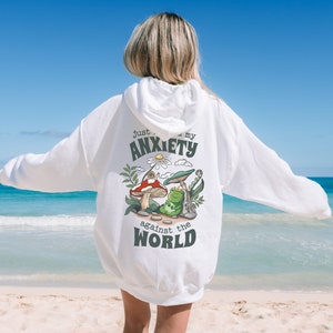 May include: White hoodie with a graphic of a frog and mushrooms. The text on the hoodie reads "Just my anxiety against the world."