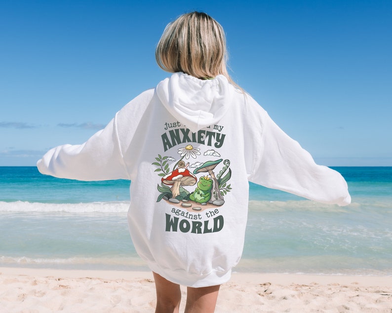 May include: White hoodie with a graphic of a frog and mushrooms. The text on the hoodie reads "Just my anxiety against the world."