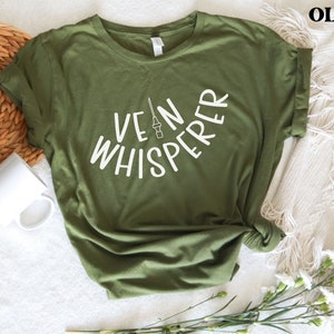以下が含まれることがあります： オリーブグリーンのTシャツに、白い文字で「Vein Whisperer」と書いてあり、スタイリッシュな針の画像が描かれています。