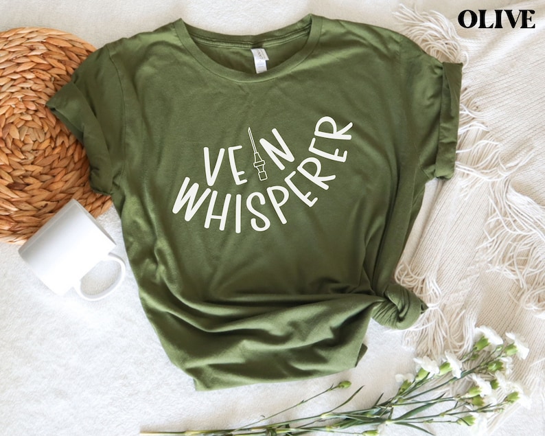 以下が含まれることがあります： オリーブグリーンのTシャツに、白い文字で「Vein Whisperer」と書いてあり、スタイリッシュな針の画像が描かれています。