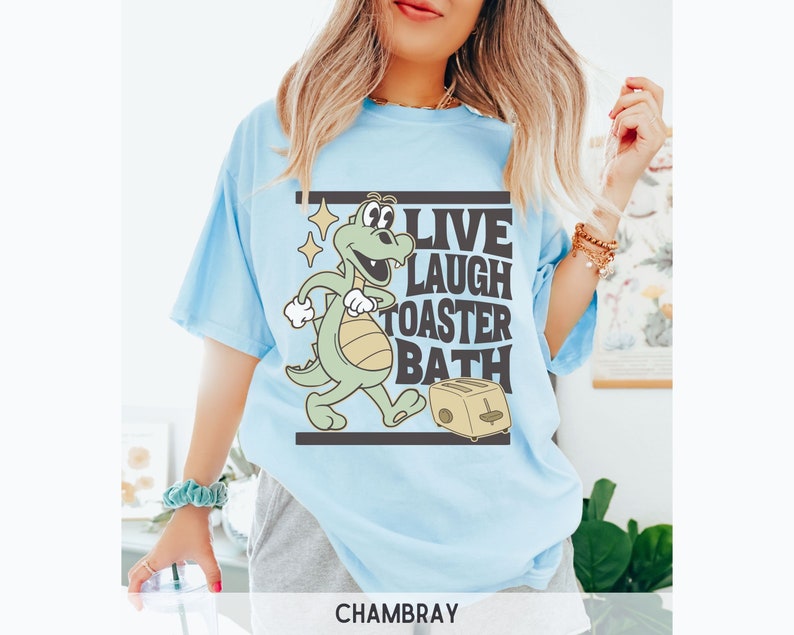 May include: Light blue t-shirt with the text "LIVE LAUGH TOASTER BATH" and a cartoon alligator with a toaster. The shirt is a chambray color.