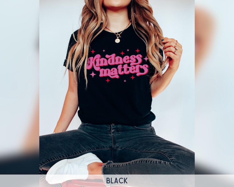 Pode incluir: T-shirt preto com a frase "Kindness Matters" em letras rosa e vermelhas, adornada com estrelas. O texto usa uma fonte retr&ocirc;. A parte superior &eacute; de estilo gola redonda.