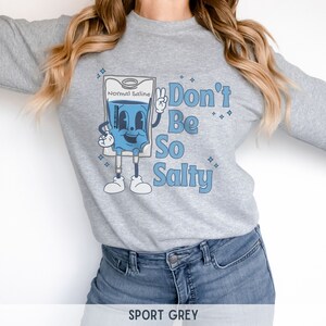 Op de afbeelding: Een grijs sweatshirt met een cartoonillustratie van een infuuszak met de tekst "Normal Saline" en de zin "Don't Be So Salty".