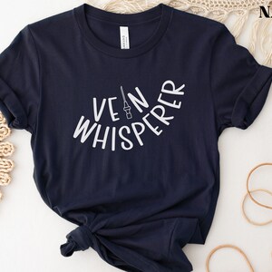 以下が含まれることがあります： 白い文字で「Vein Whisperer」と書いてあるネイビーブルーのTシャツと、小さな針の画像です。