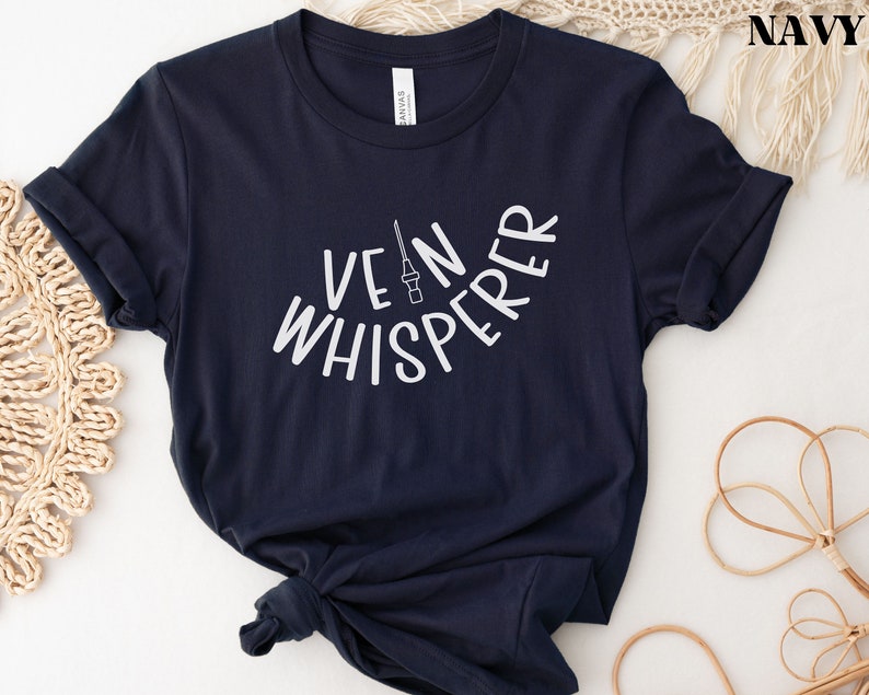 以下が含まれることがあります： 白い文字で「Vein Whisperer」と書いてあるネイビーブルーのTシャツと、小さな針の画像です。