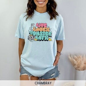 Pu&ograve; includere: Una maglietta blu chiaro con una stampa grafica del testo "Live Laugh Toaster Bath" in lettere a bolle colorate. La grafica include anche una doccia, un tostapane, un'anatra di gomma e bolle di sapone.