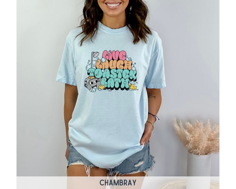 Pu&ograve; includere: Una maglietta blu chiaro con una stampa grafica del testo "Live Laugh Toaster Bath" in lettere a bolle colorate. La grafica include anche una doccia, un tostapane, un'anatra di gomma e bolle di sapone.