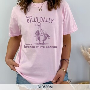 May include: Pink t-shirt with a graphic of a duck wearing a doctor's coat and stethoscope. The text on the shirt reads "born to dilly dally forced to update white boards".