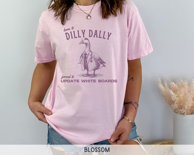 May include: Pink t-shirt with a graphic of a duck wearing a doctor's coat and stethoscope. The text on the shirt reads "born to dilly dally forced to update white boards".