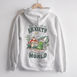 May include: Gray hooded sweatshirt with a green frog, mushroom, and snail graphic. The text "Just me and my anxiety against the world" is printed on the back.